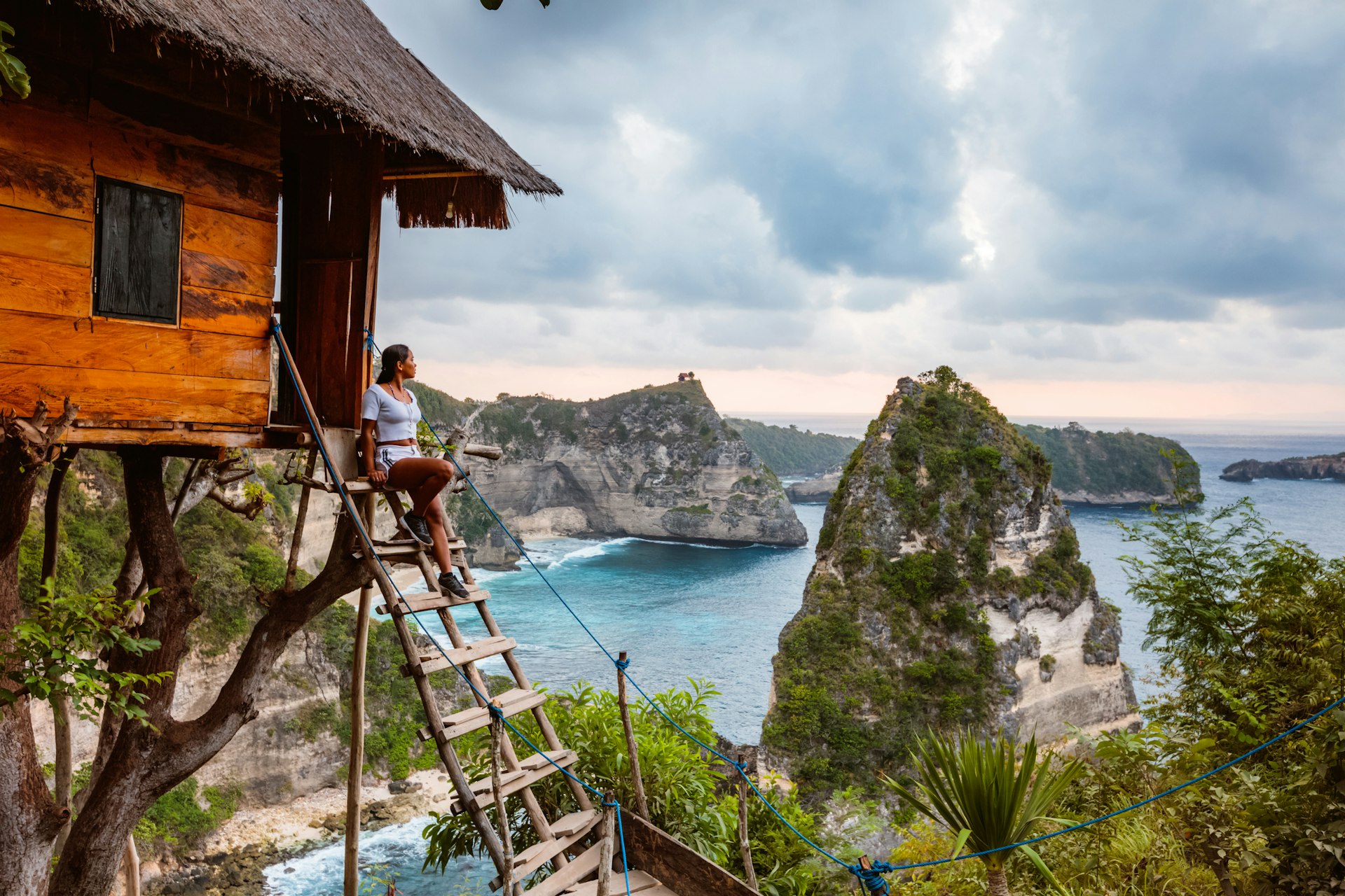 A solo woman sits at the top of a ladder leading up to a treehouse overlooking a bay with several rocky islets