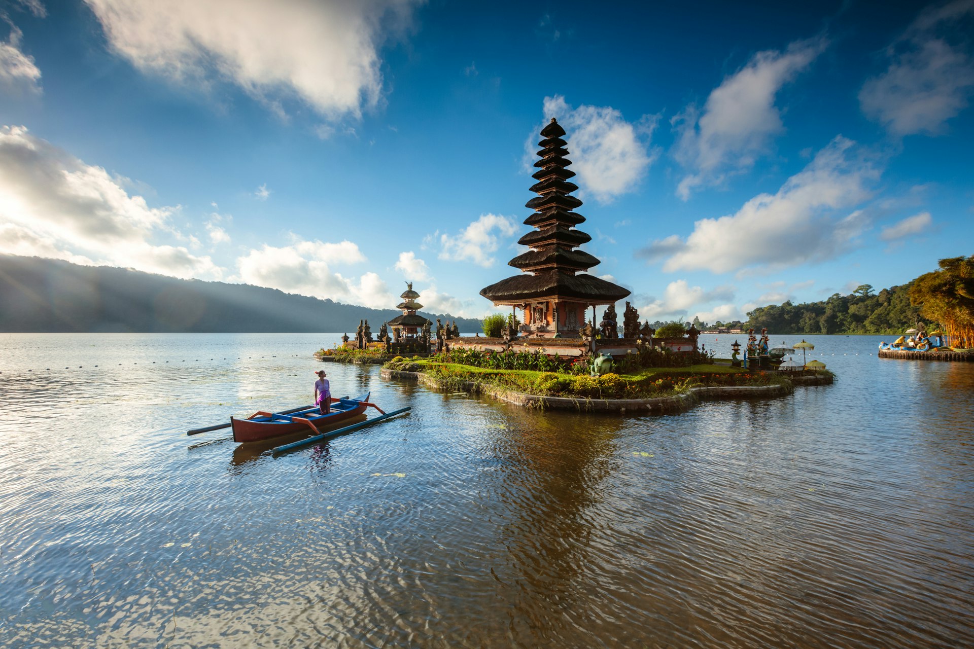 A Balinese woman rows a boat towards the Ulun Danu Beratan temple, Bedugul in Bali, Indonesia with blue skies and fluffy clouds above