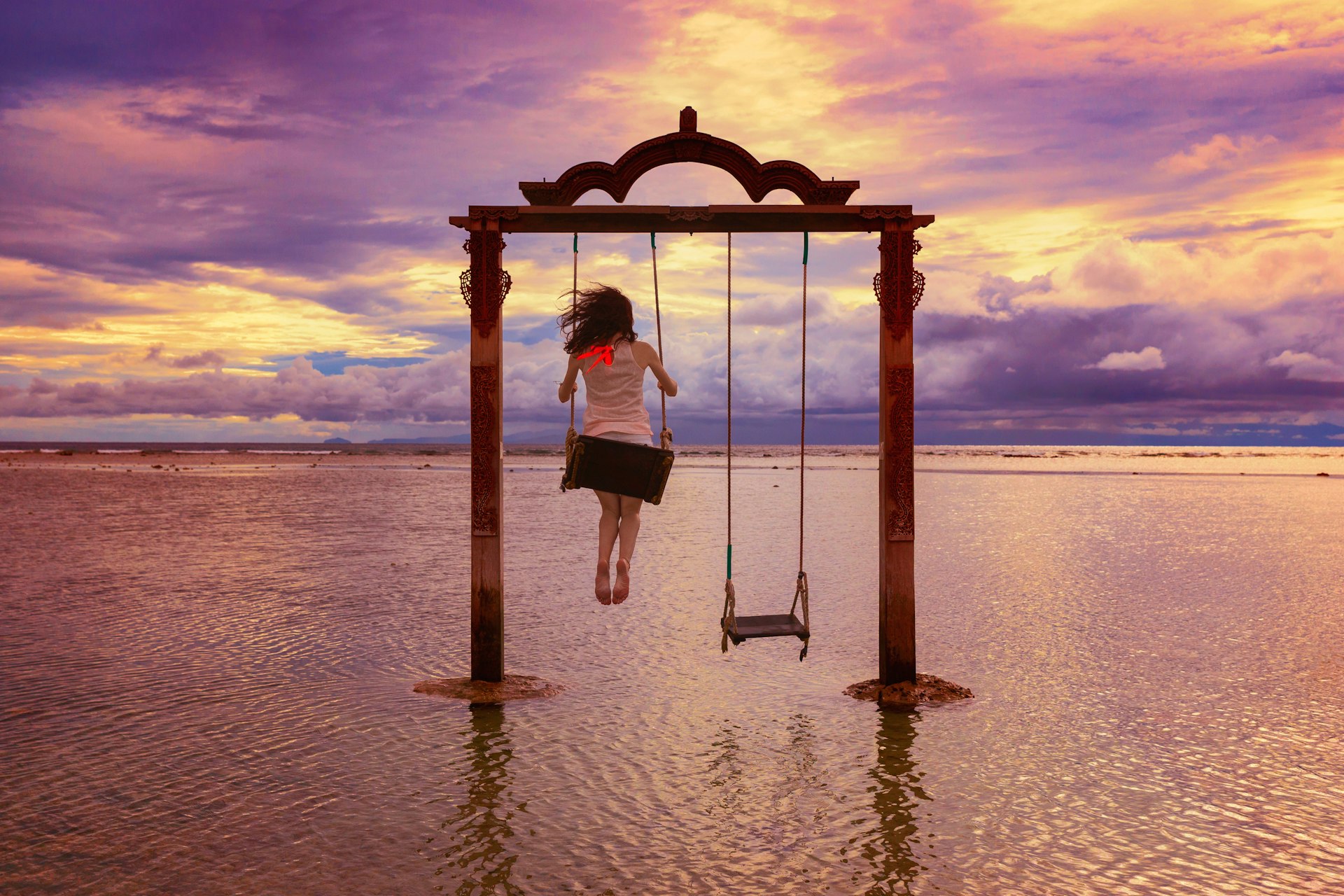 A woman swings high on a swing set in the shallows of the ocean. The sun is setting casting a pink hue across the sky