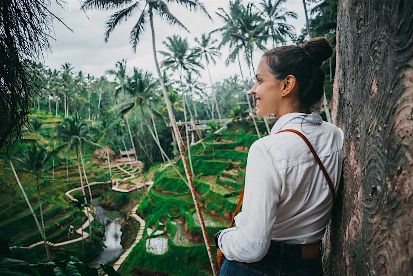 Seorang wanita muda kulit putih dengan rambut cokelat dengan kemeja putih melihat ke sawah dan pohon palem di Ubud, Bali 