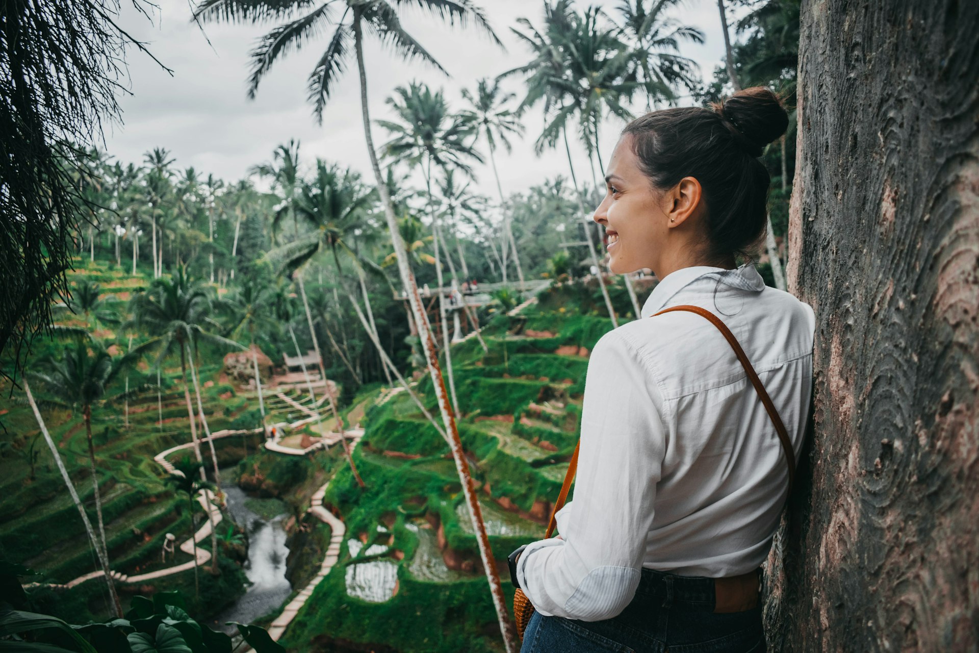 A young white woman with brown hair in a white shirt looks over the rice terraces and palm trees of Ubud, Bali 