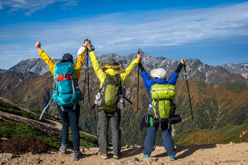 Three hikers look triumphantly at a mountain range with their arms raised