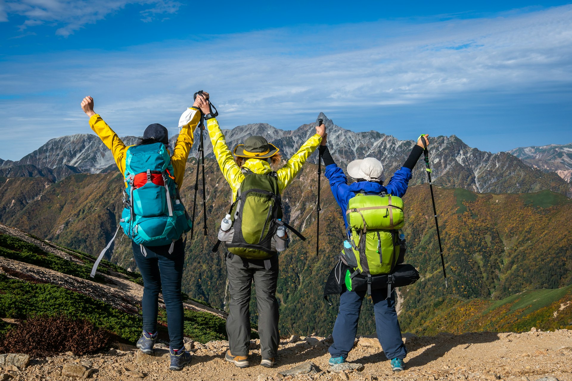 Three hikers with their arms in the air in a triumphant stance face out towards a mountain range