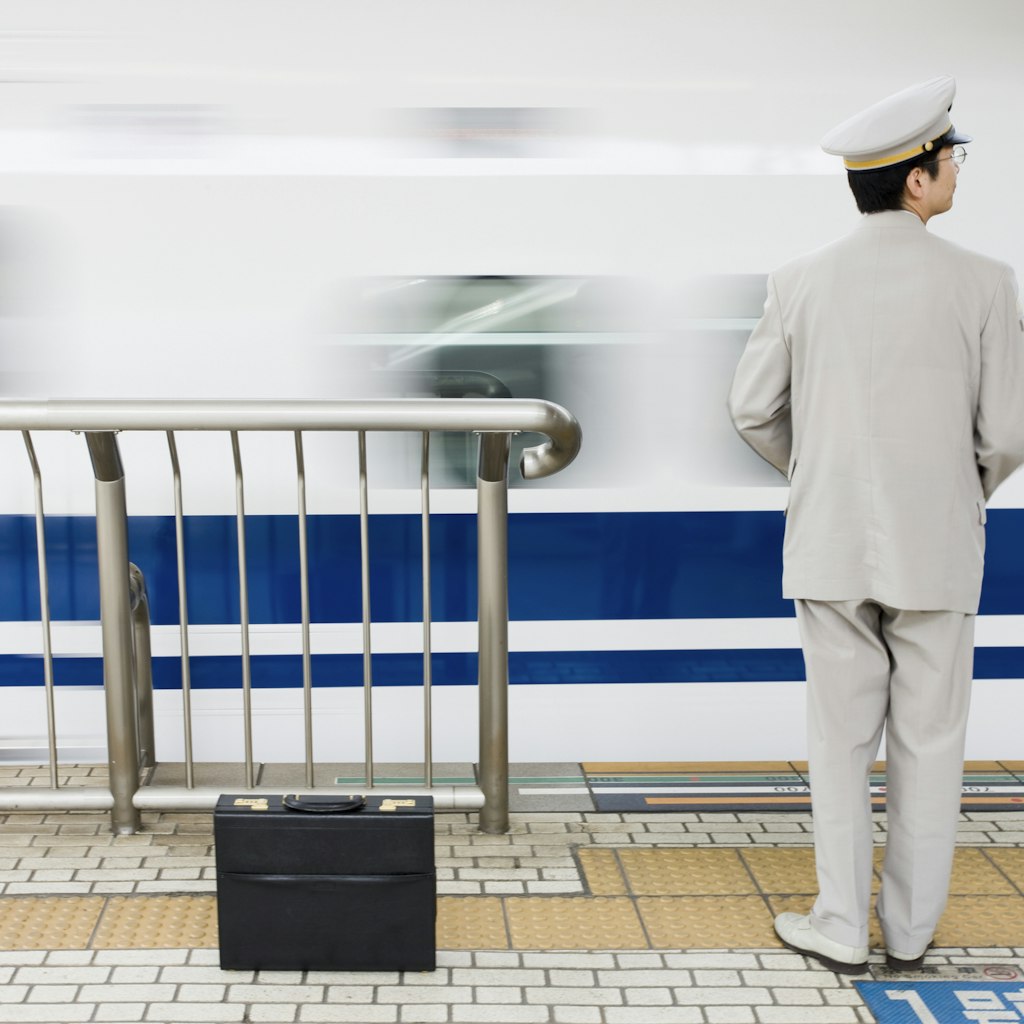 A smartly dressed Nozomi bullet train worker in all white suit and cap with black briefcase watches the train come into the platform in Kyoto, Japan