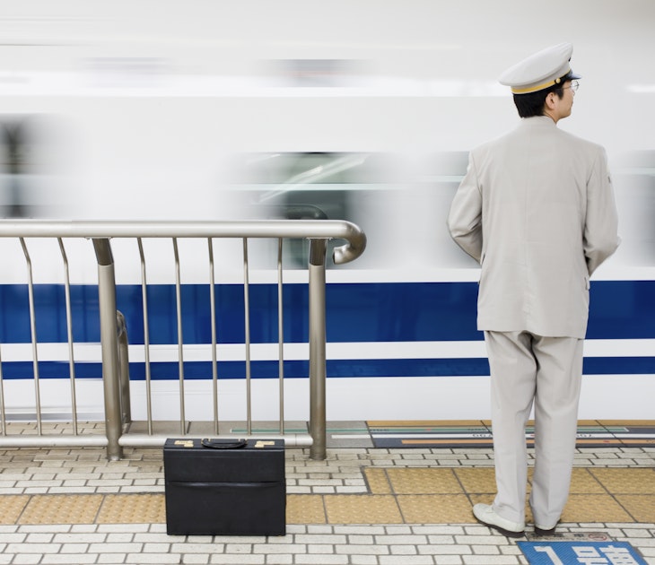 A smartly dressed Nozomi bullet train worker in all white suit and cap with black briefcase watches the train come into the platform in Kyoto, Japan