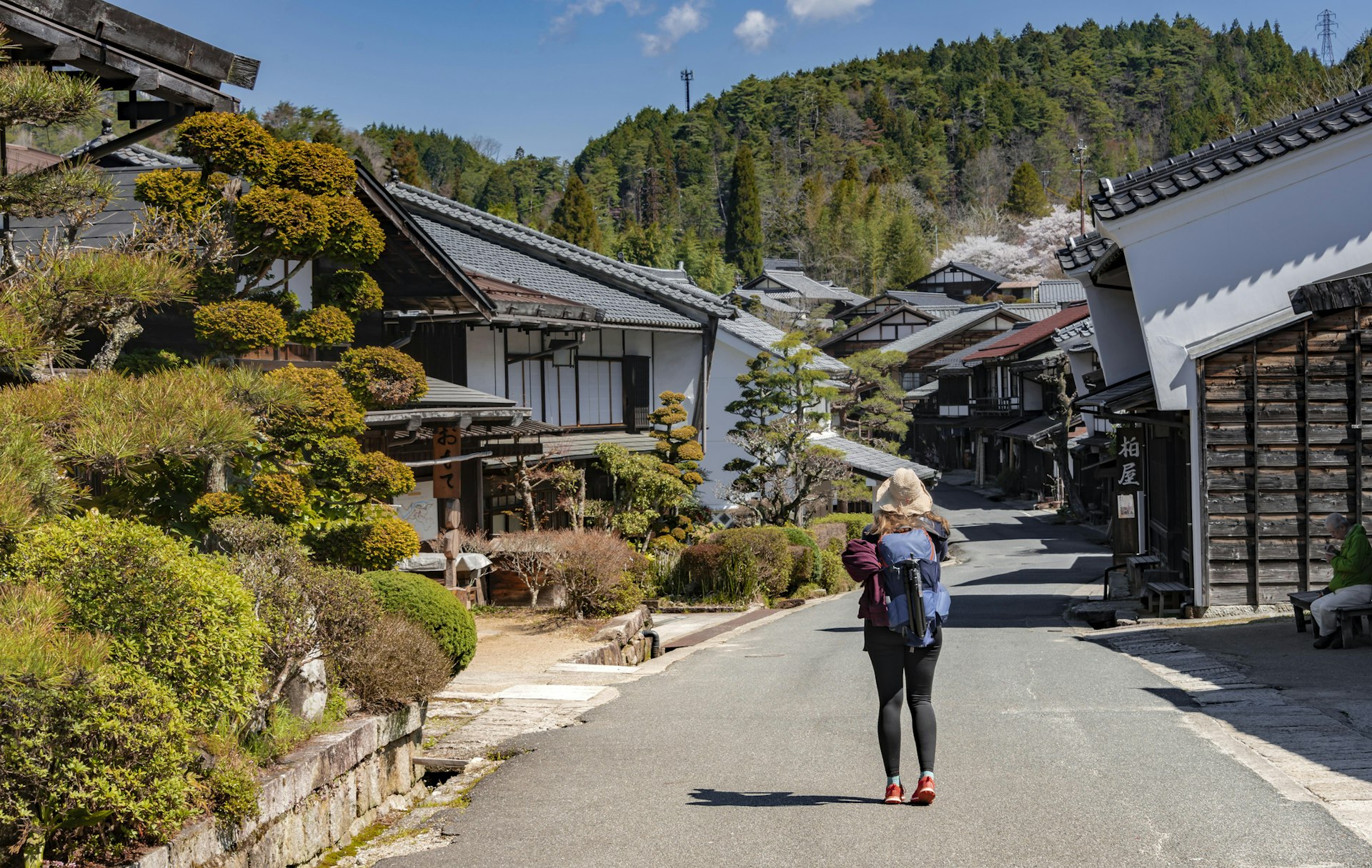 A solo hiker on the Nakasendo path through a traditional Japanese village. The path is lined with wooden low-rise houses