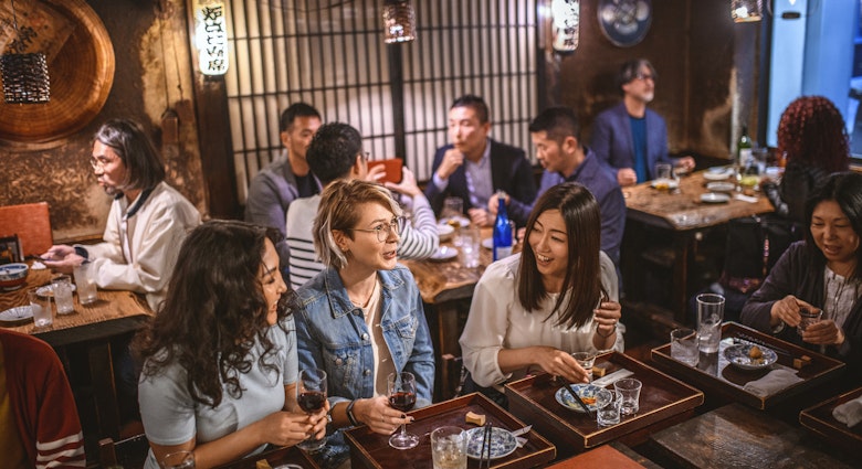Wide angle view of Mongolian, Japanese, and Caucasian women sitting at sushi bar and other patrons in Tokyo izakaya.