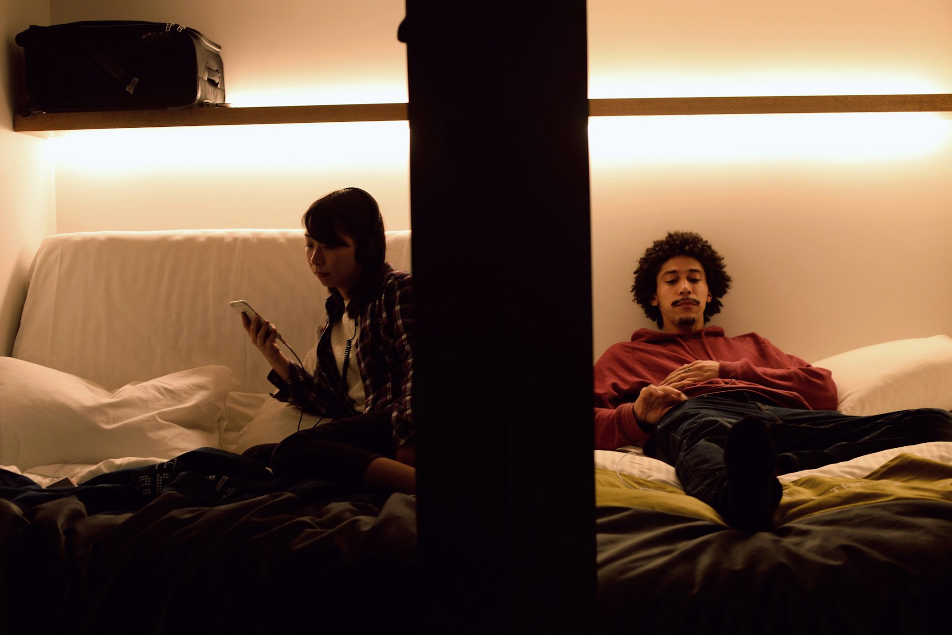 Two people sat in their capsule rooms in a capsule hotel. The bed fills the space with a shelf above to put belongings on