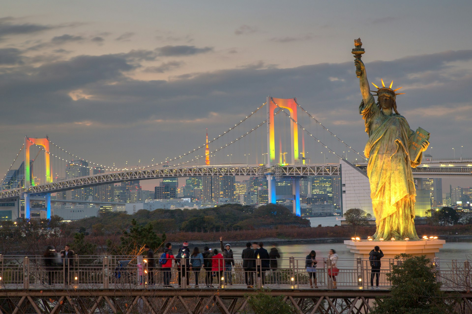 People stand on a platform admiring a suspension bridge in the background it in rainbow colors. A replica Statue of Liberty stands in the foreground