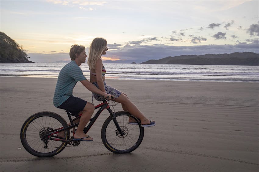 A man steers a bike while a young woman sits on top of the handlebars as they bike on Playa Venao in Panama. 