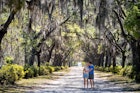 A man kisses a woman's kiss on a path flanked with Spanish moss trees in Savannah. 