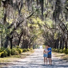 A man kisses a woman's kiss on a path flanked with Spanish moss trees in Savannah. 