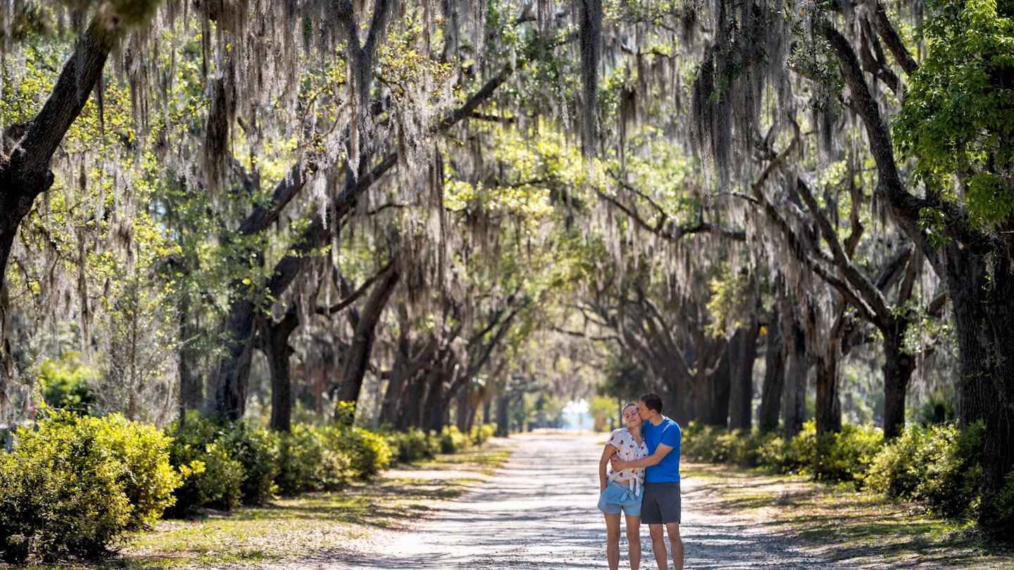 A man kisses a woman's kiss on a path flanked with Spanish moss trees in Savannah. 