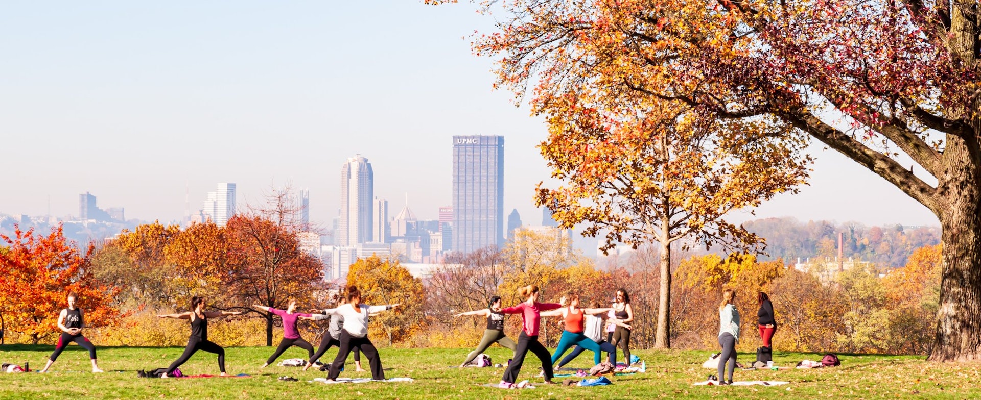 A group of women participate in a yoga class in Schenley Park with downtown Pittsburgh in the background on a hazy fall morning 