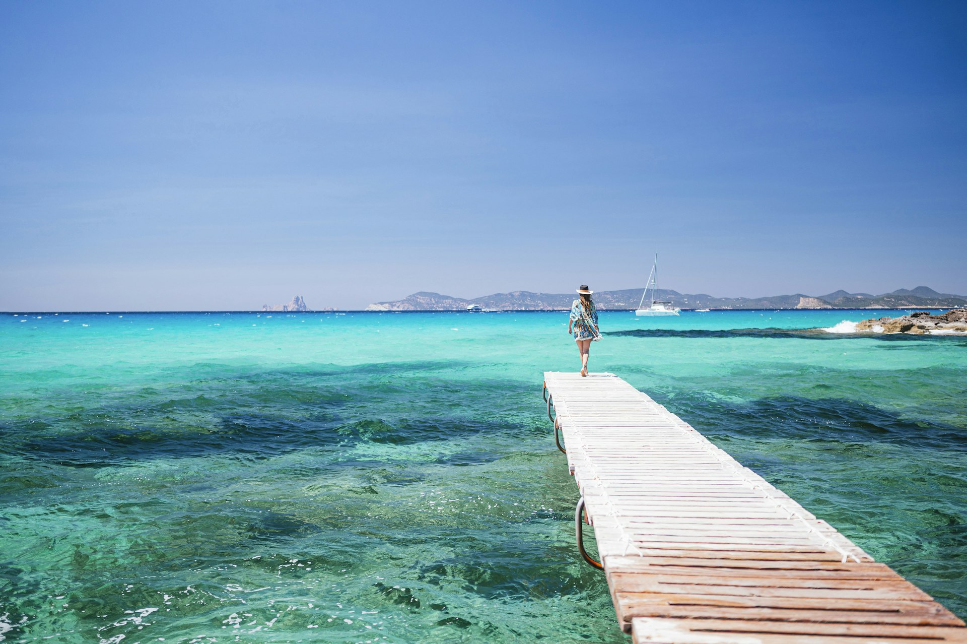 A woman walks along a wooden pontoon towards a turquoise ocean on a beautiful sunny day. There's a boat in the distance and an island beyond