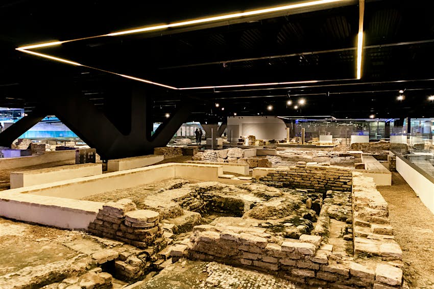 Un muzeu arheologic cu ruine aliniate la pământ și vizitatori care privesc de pe alei