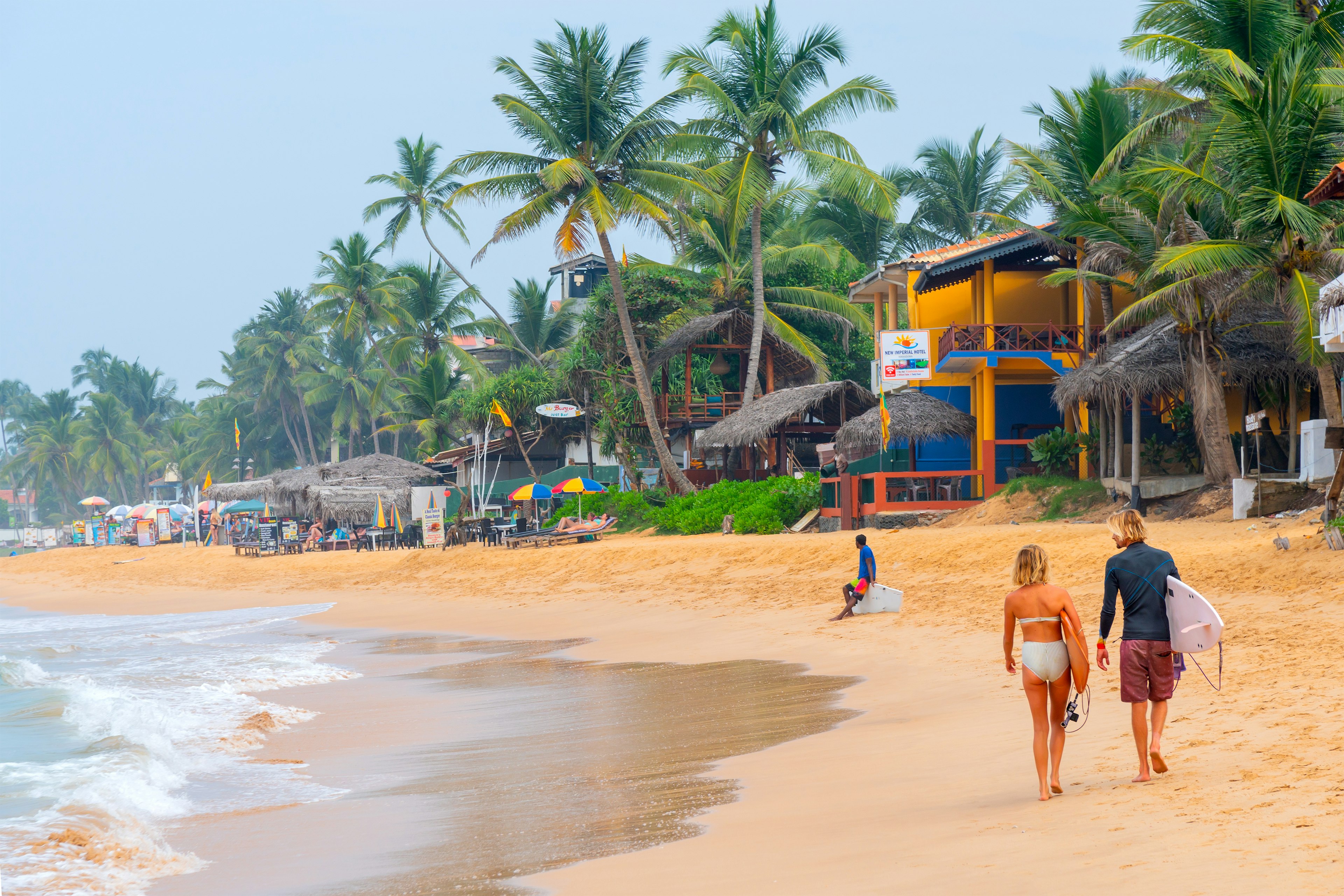 A woman and a man carrying surfboards walk along a beach lined with bars, beach shacks, and palm trees