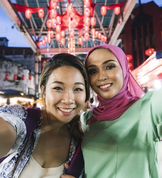Two friends, one chinese young woman and one muslim mid adult woman,  having a selfie at the market together