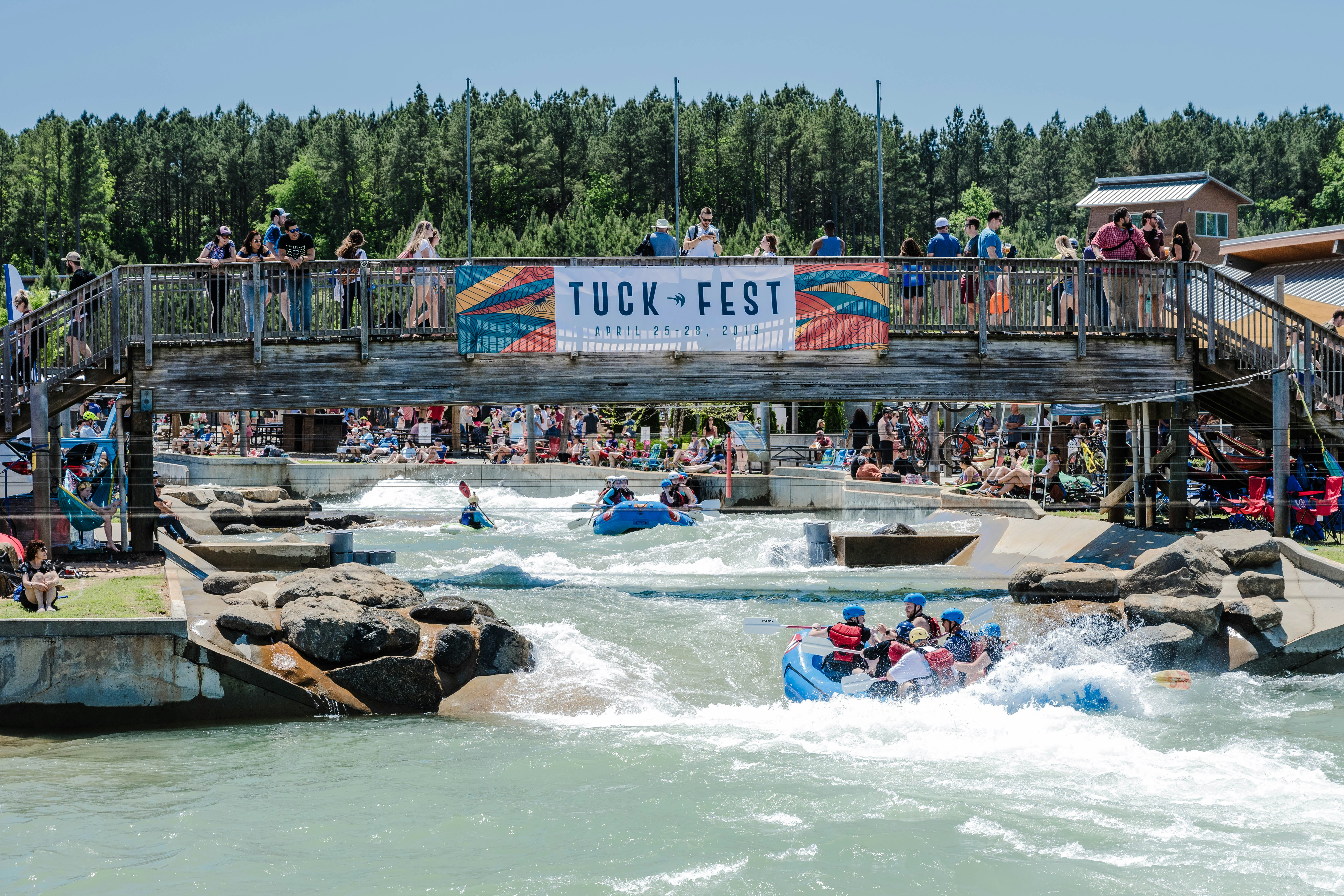 A group of people in a large rubber raft paddle on rushing white water at the US National Whitewater Center in Charlotte, NC. There is a large sign that says "Tuck Fest" hanging from a wooden bridge above the water.