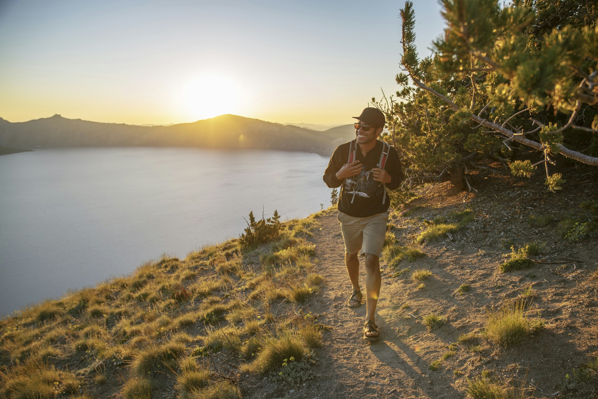 A smiling black hiker in cap and sunglasses walks along the shoreline of Crater Lake in Oregon, USA with a camera around his neck as the sun begins to set over the hills in the background