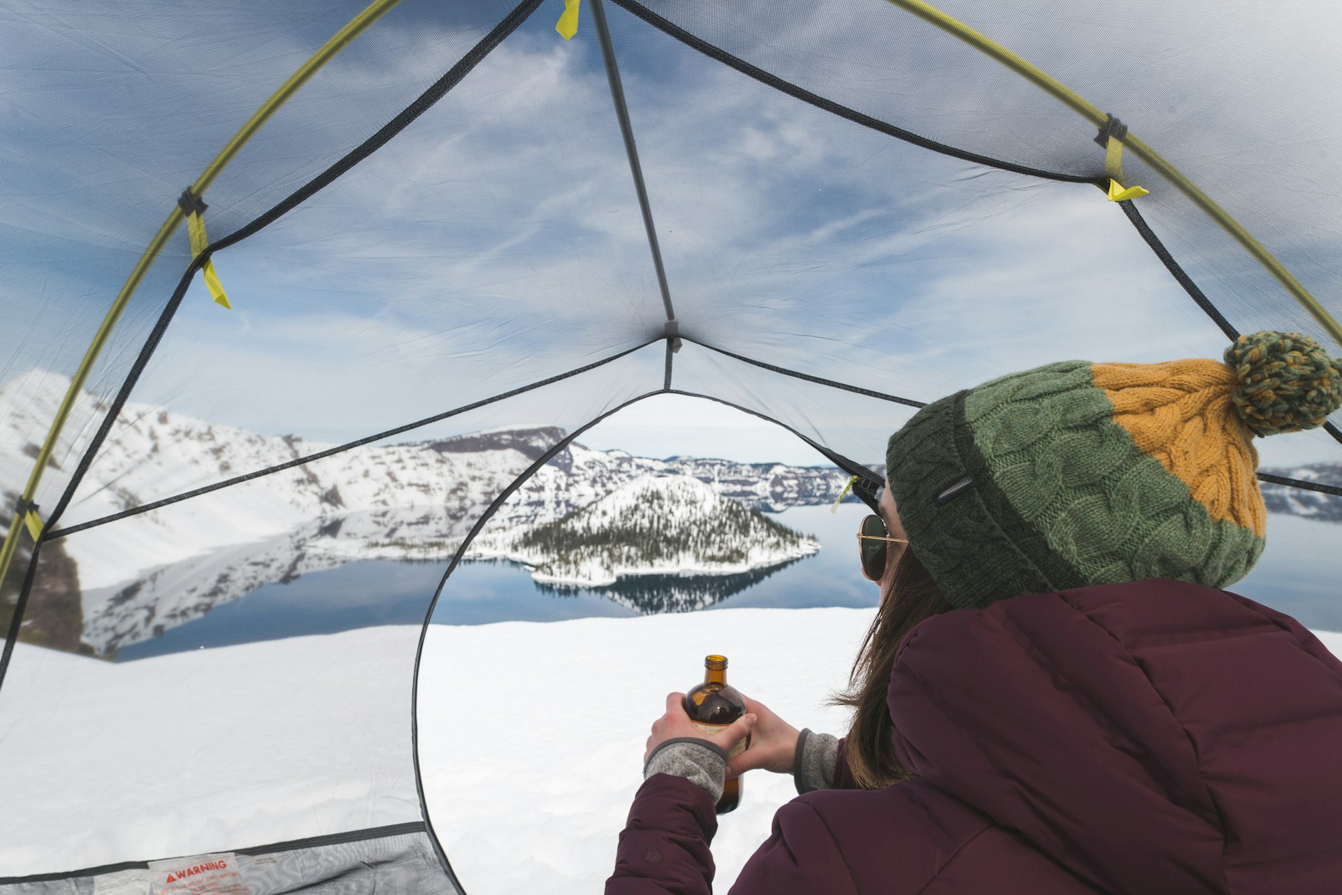 A young female hiker relaxes in her tent in the snow overlooking a Crater Lake and drinks a cold beverage. The shot is from behind her.