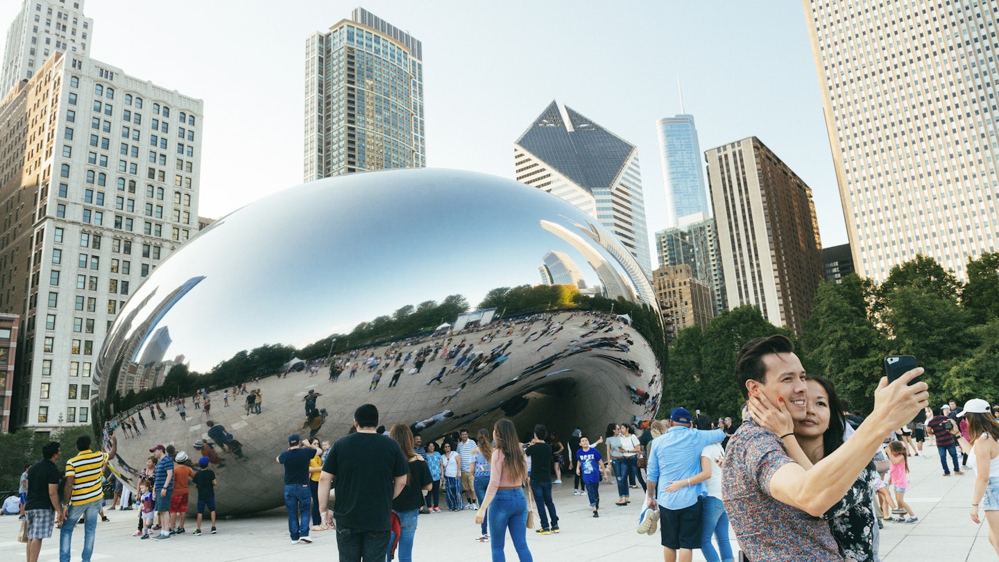 This is a horizontal color photograph of people crowded around the landmark downtown Chicago's Cloudgate in Millennial Park. The urban cityscape is reflected in the mirrored surface. A couple takes a selfie in the foreground.