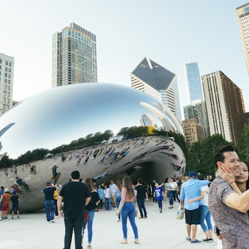 This is a horizontal color photograph of people crowded around the landmark downtown Chicago's Cloudgate in Millennial Park. The urban cityscape is reflected in the mirrored surface. A couple takes a selfie in the foreground.