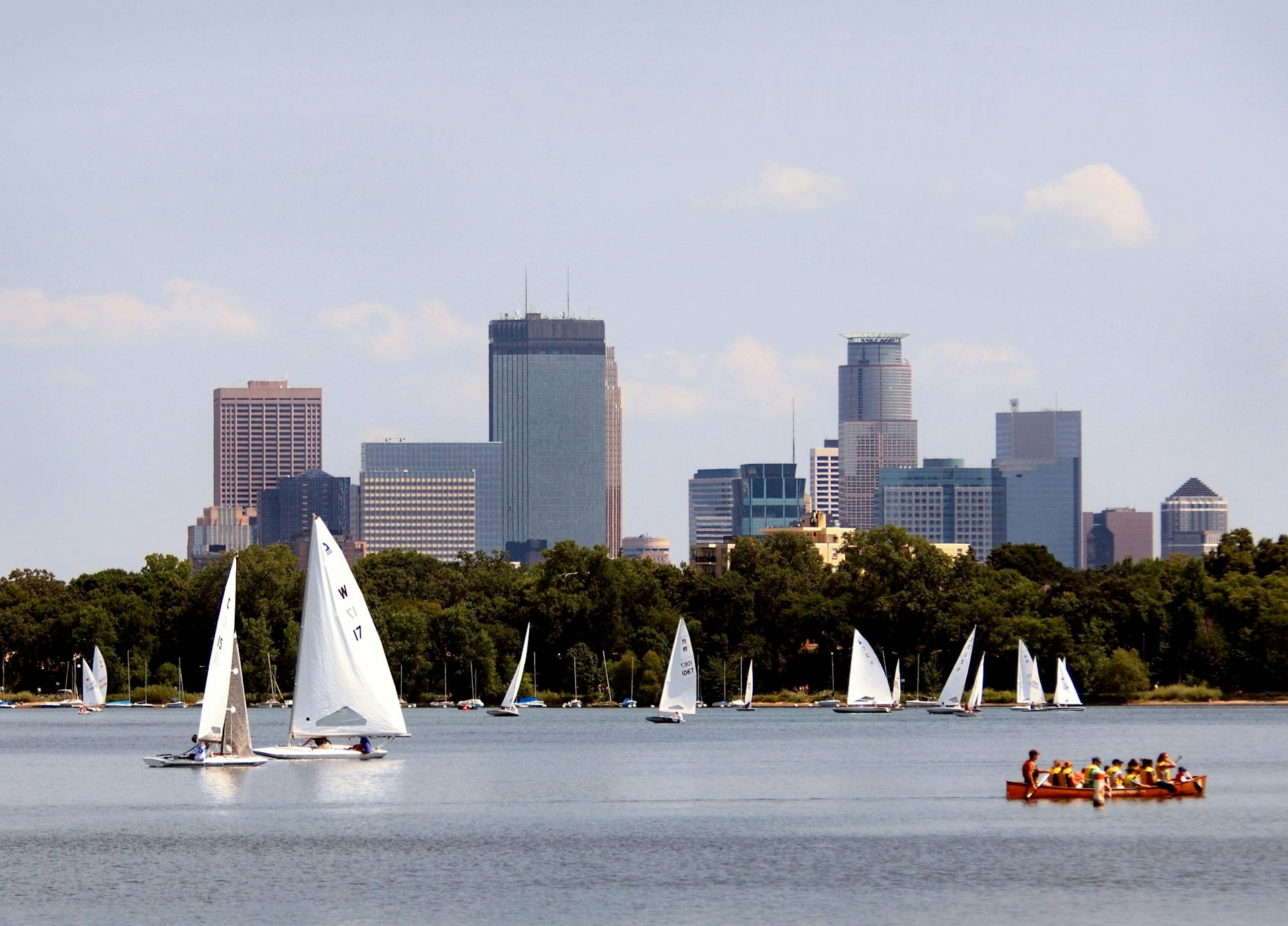Sailboats with white sails on a lake at the edge of a city. There are several tall buildings in the background