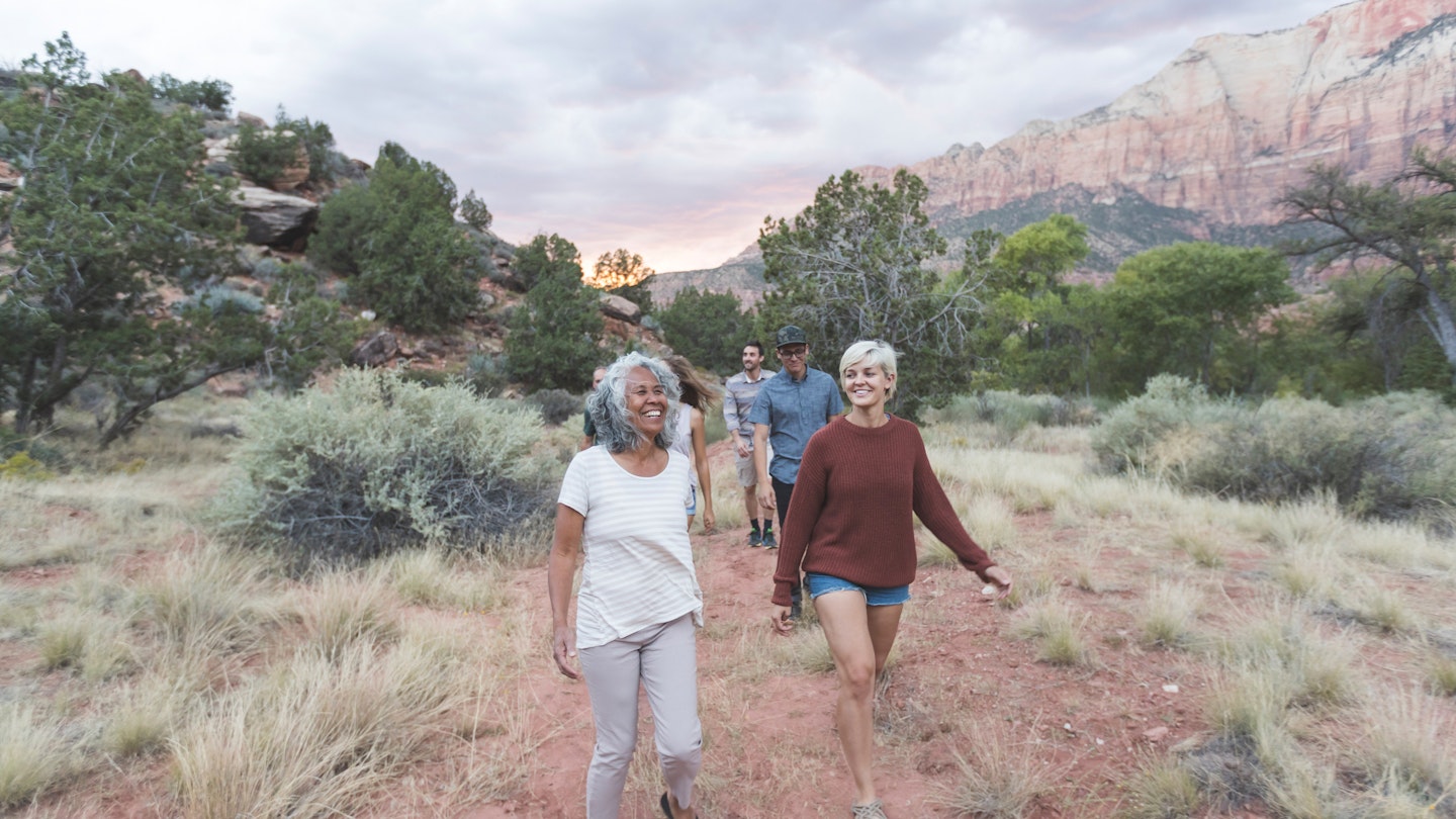 A multiethnic group of friends walk through the Utah desert. Two females are in front - one in her 20s, one in her early 60s.