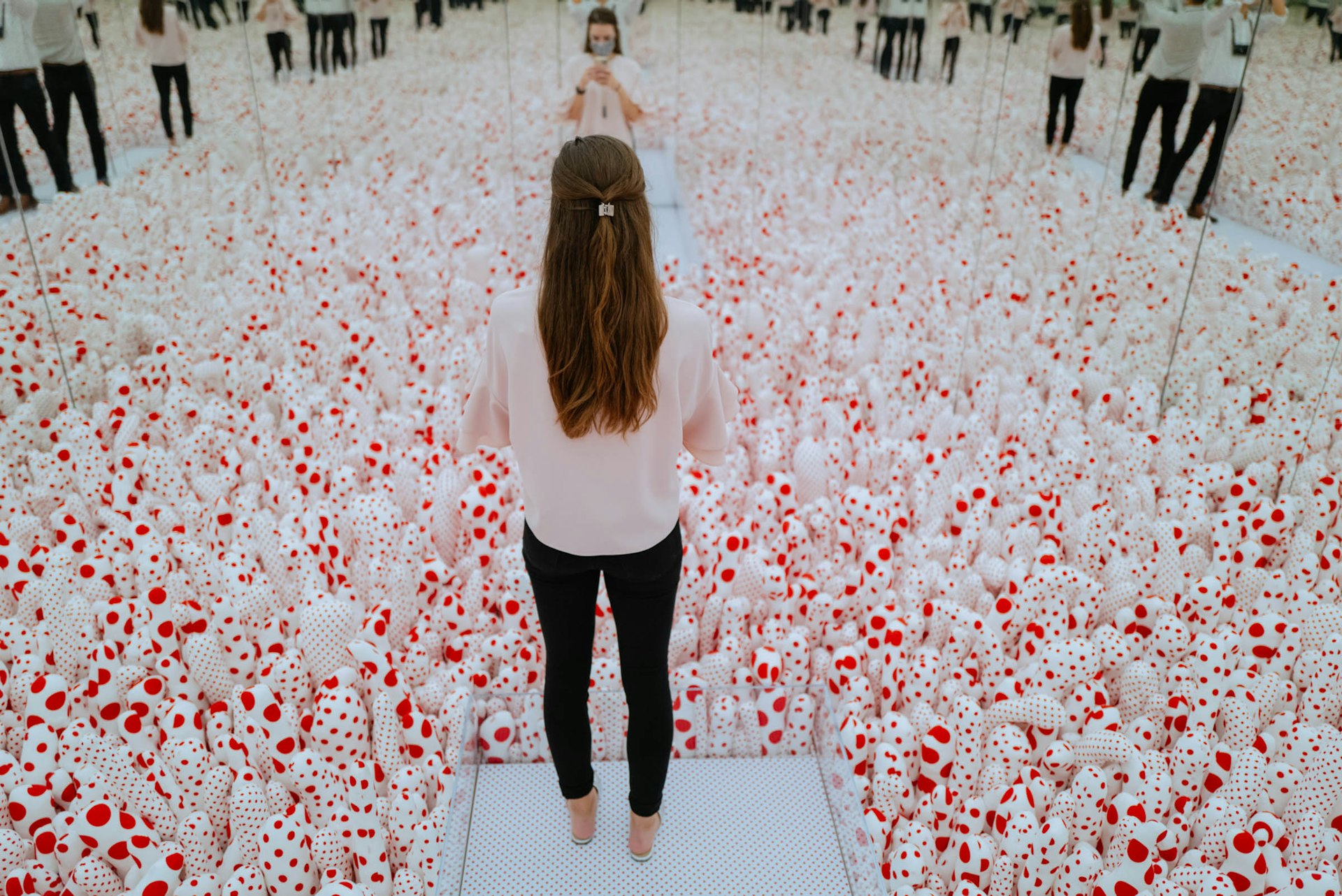 A visitor standing in Yayoi Kusama’s Infinity Mirror Room—Phalli’s Field (1965/2017), a mirror-lined room filled with phallic red-and-white polka-dotted sculptures