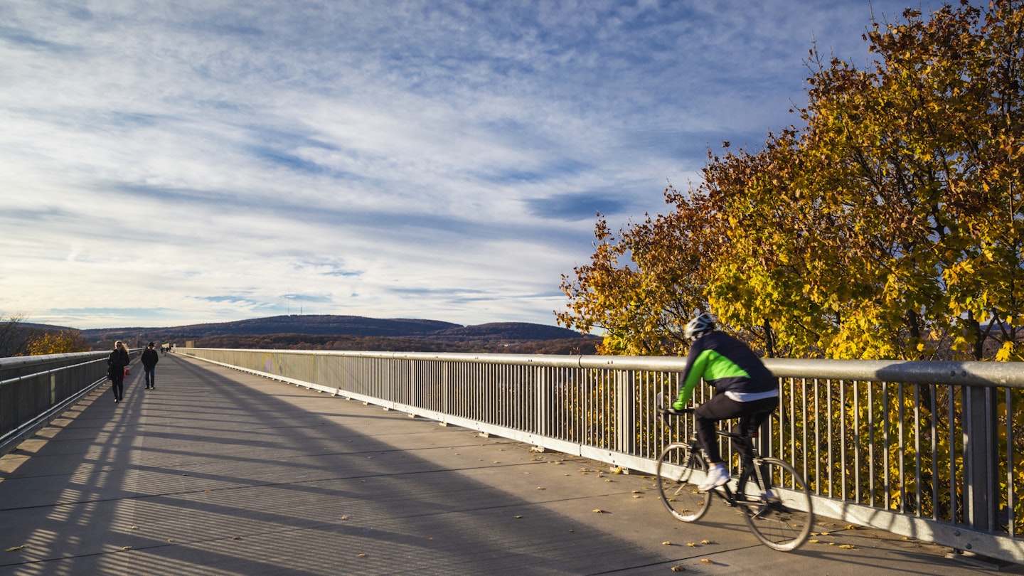 A cyclist bikes across the Walkway Over the Hudson in Hudson NY. There's a couple walking in the opposite direction. 