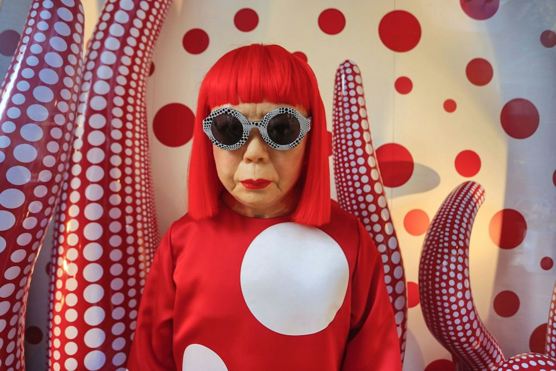A portrait of Yayoi Kusama, with red hair and googles, in front of her signature polka dot art. 