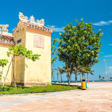 Da Nang, Vietnam - May 7, 2018: a tiny old temple on the seafront sidewalk with the distant view of palm trees, several walking people and sea against blue sky on My Khe Beach.