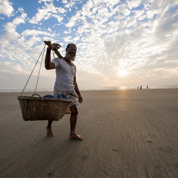 "Cox's Bazar, Bangladesh - February 10, 2013: A man is carrying seafood in barrel on shoulder walking on the beach of Cox's Bazar, Bangladesh."