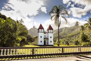 A DSLR photo of Haapiti Chatolic Church in Moorea Island, French Polynesia. The church is located on a big green lawn surrounded by dense tropical forest and mountains in the background. It is a bright sunshine day with blue sky and scattered white clouds. There is no one around.