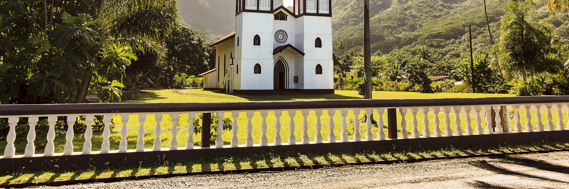A DSLR photo of Haapiti Chatolic Church in Moorea Island, French Polynesia. The church is located on a big green lawn surrounded by dense tropical forest and mountains in the background. It is a bright sunshine day with blue sky and scattered white clouds. There is no one around.
