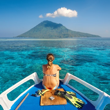 Young woman in swimsuit sit on boat with a mask and flippers looking to a clean sea and volcano Manado Tua. North Sulawesi, Indonesia.