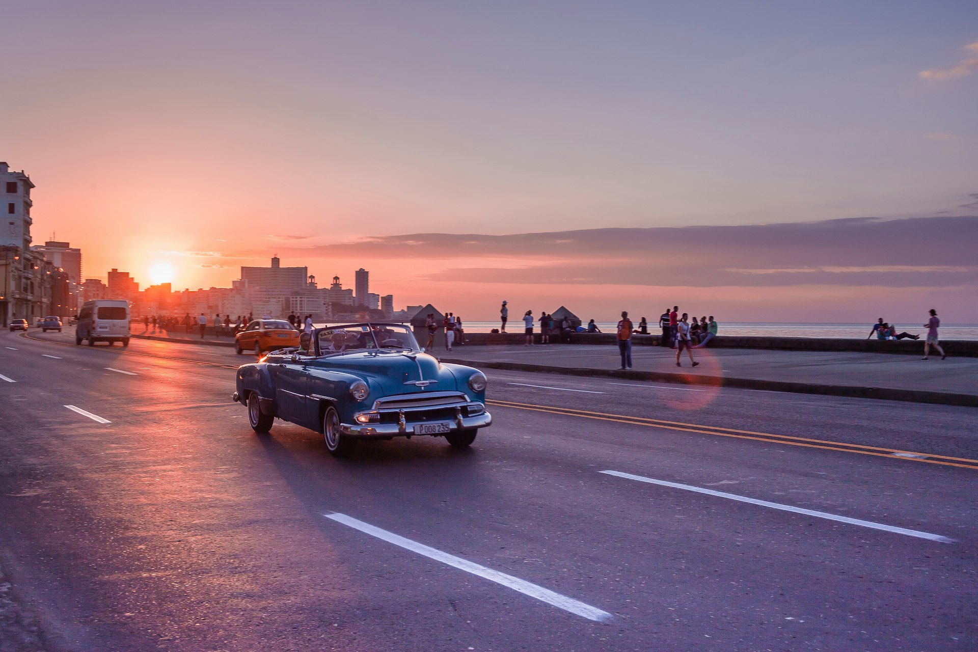 A classic car drives along a seaside road in Havana as the sun sets