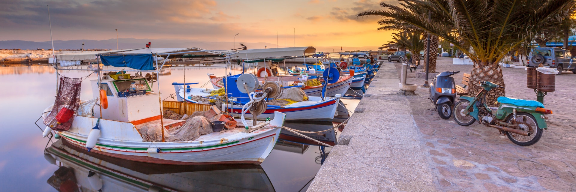Fishing harbor scene in Greece with boats, palm trees and scooters at sunrise on a beautiful tranquil summer day