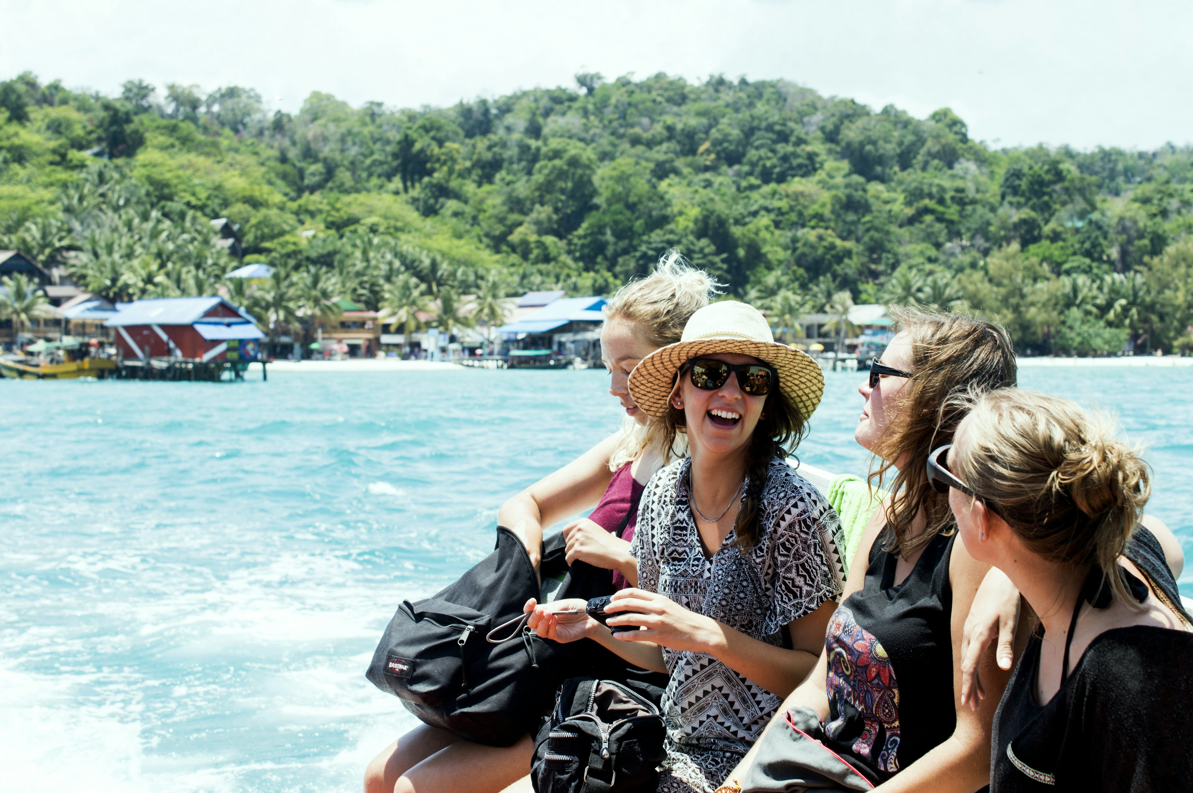 A group of female friends laugh and smile as the boat they're on approaches an island lined with huts and palm trees