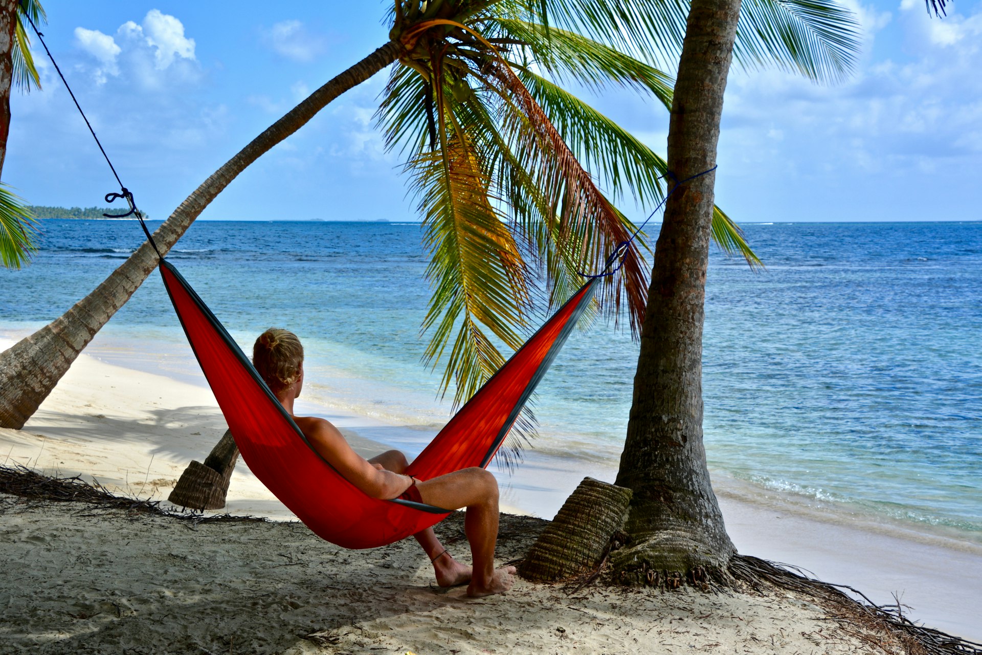 A man in a bathing suit looks out at the ocean while relaxing in a hammock strung between palm trees on Relaxing in a hammock on Islas San Blas in Panama