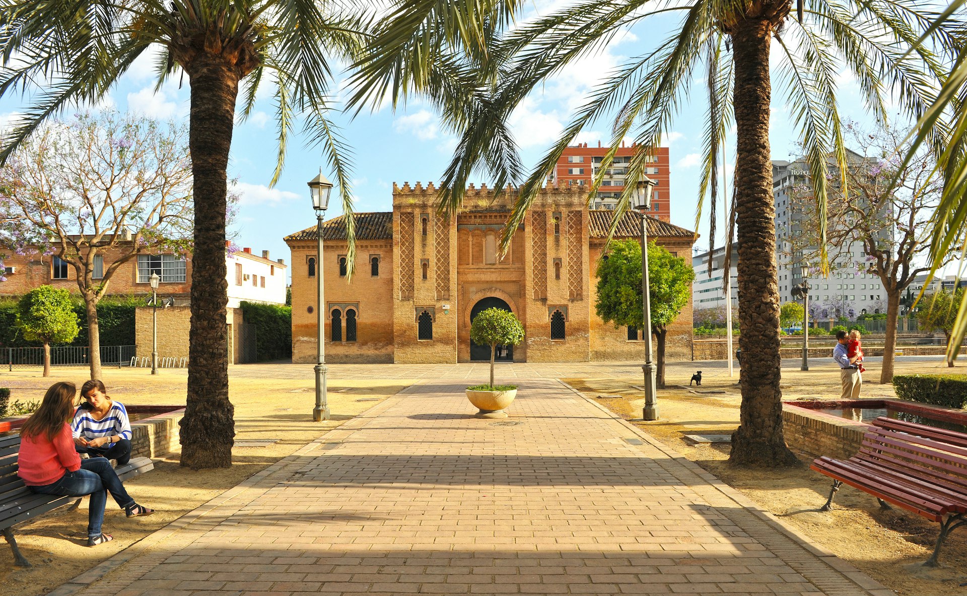 People on benches under date palms in front of a historic palace at Jardines de la Buhaira, Sevilla, España