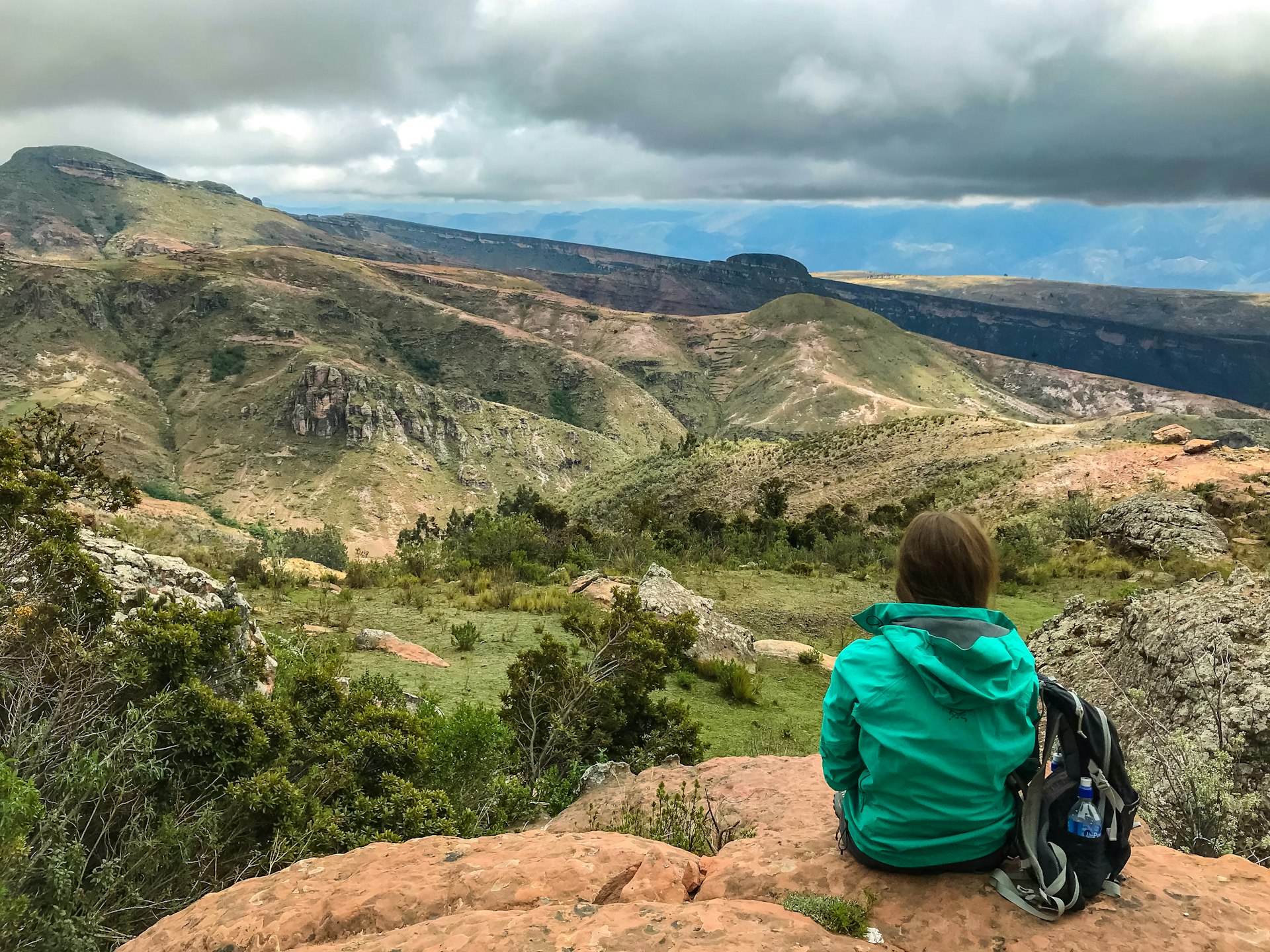 Woman with a green raincoat shot from behind, sitting at the edge of a rock point looking at the landscape at Torotoro National Park, Potosí, Bolivia