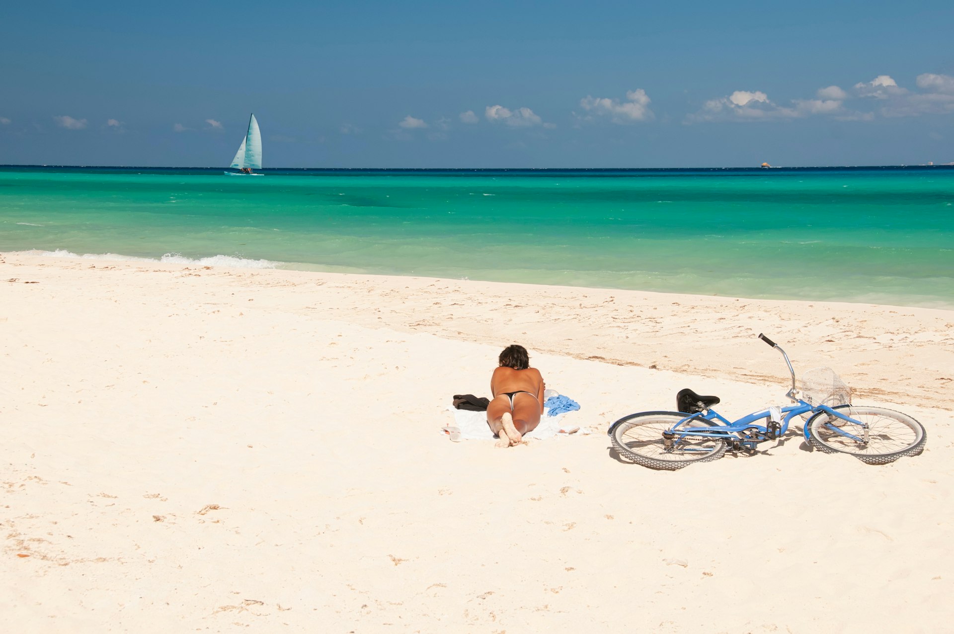 A young woman lies on the sand in a bikini, reading, next to her bicycle with a sailboat on the Caribbean Sea beyond
