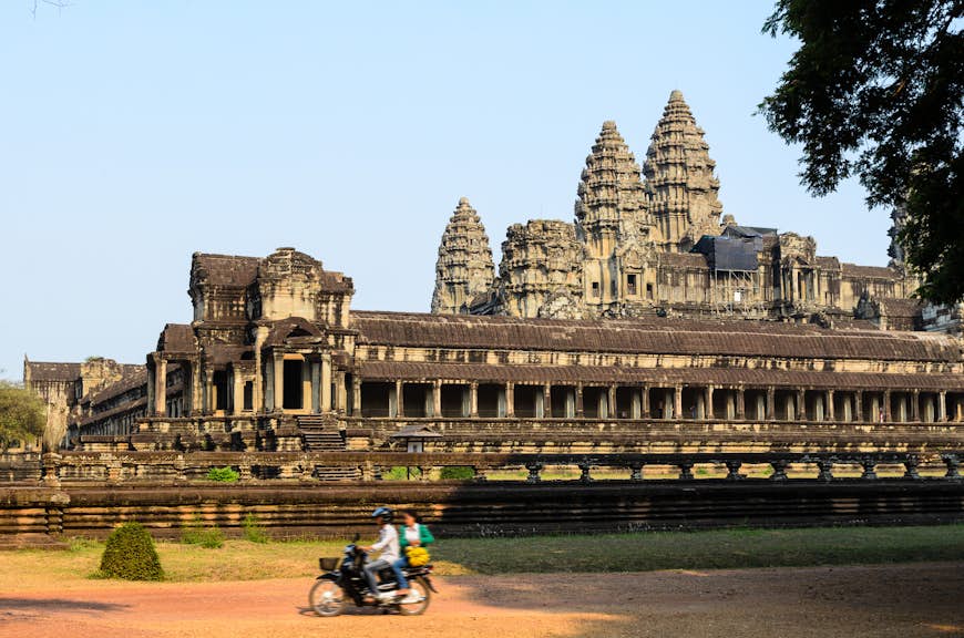 A motorbike with two people on it passes on a dirt road by a temple at Angkor Wat, Siem Reap, Cambodia, Southeast Asia