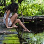 Woman with curly hair sitting on bridge stretches out leg towards water in green forest in Bocas del Toro Islands, Panama.
