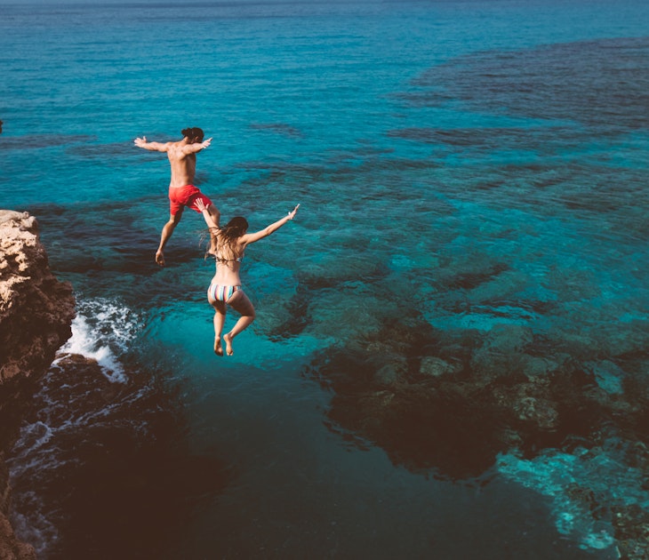 Young active man and woman diving from high cliff into tropical island blue sea water