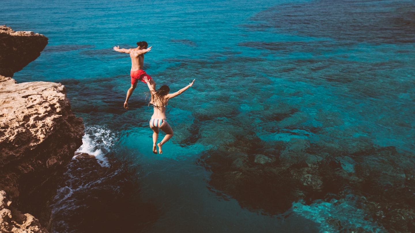 Young active man and woman diving from high cliff into tropical island blue sea water