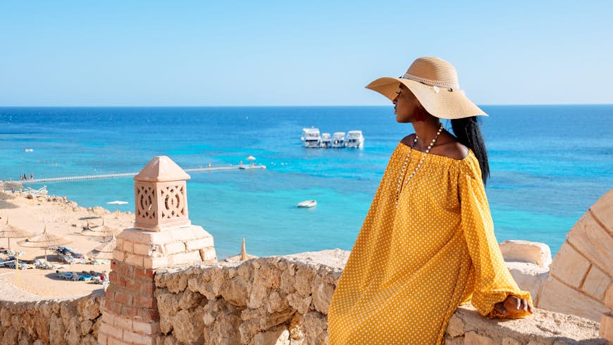 A woman wearing a yellow dress and a sunhat stands at an overlook above a sandy beach and turquoise sea in Sharm El Sheikh, Egypt
