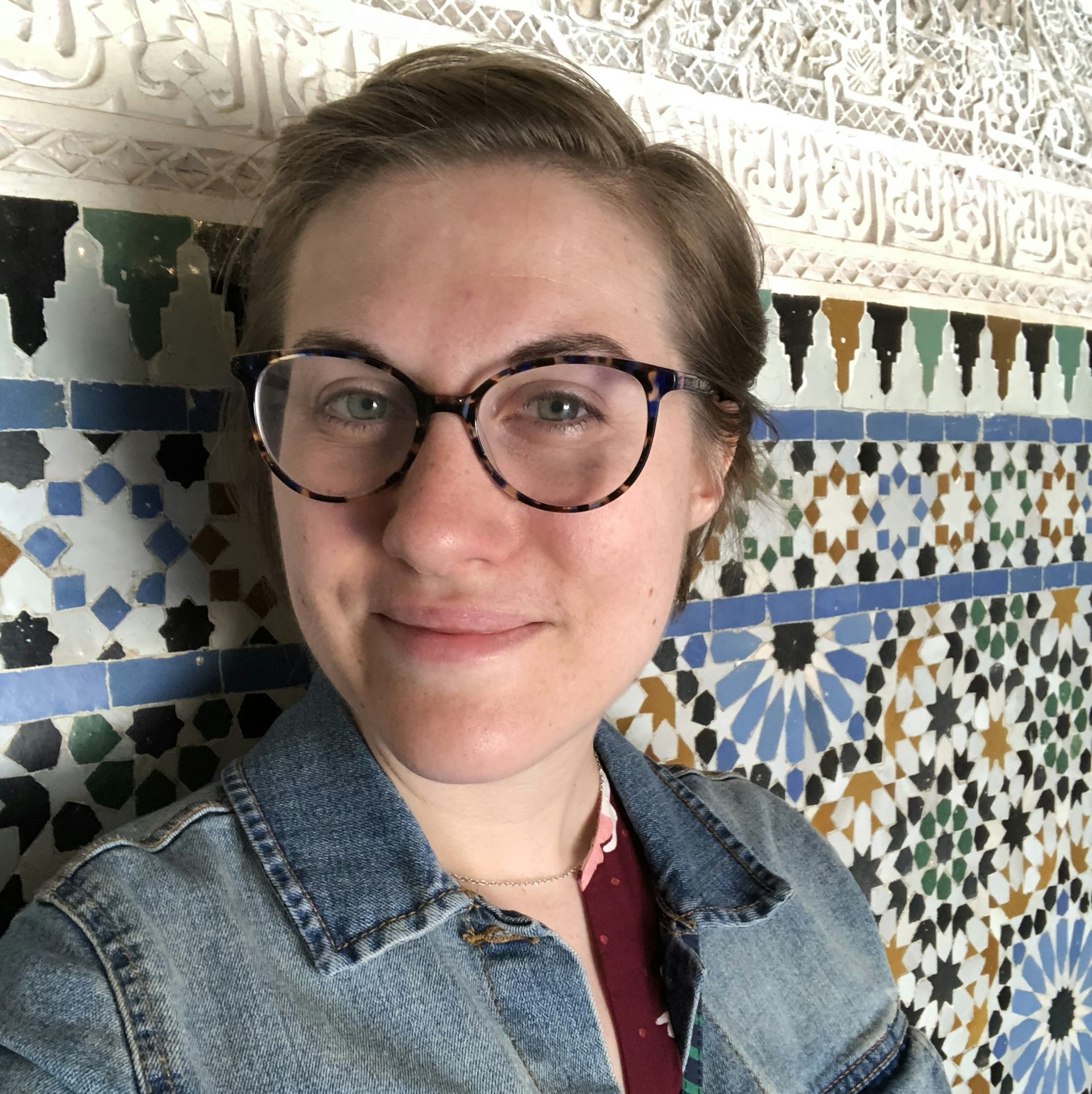 A woman wearing glasses looks into the camera in front of a tile wall. 